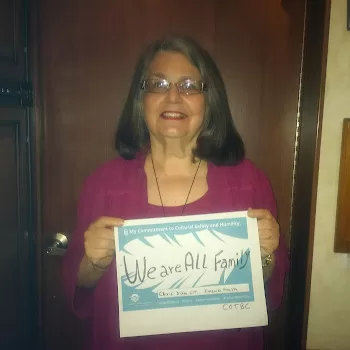 my wonderful wife older and smiling looking out holding a sign with two fingers of each hand that says in black letters We Are All Family