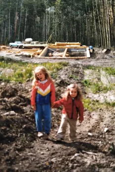 young sisters holding hands walking up hill with timber frame house foundation in background and tall trees around building site