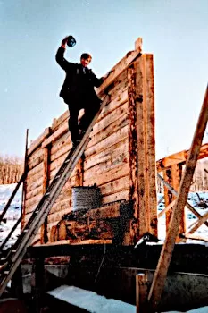 rob high up on ladder above timber wall snow behind on water dugout berm as second floor timber support posts can be seen